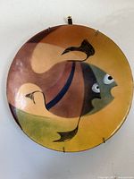 Full view of the vintage enamelled copper circular wall hanging showing the twin fish design in abstract colors including mustard yellow, green, and brown. Artist signed.