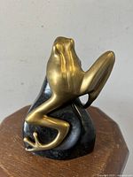 Solid brass frog sculpture sitting on black stone rock base showing front and side views, highlighting detailed feet and smooth finish.