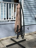 Photo of the full umbrella and black metal stand with cross-shaped base, folded and standing upright outside near a porch railing.