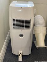 Front view of Amcor portable air conditioner with remote control placed in front of unit and exhaust hose beside it.