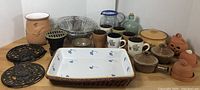 Wide shot of assorted kitchen items including mugs, ceramic pots, glass jug, and decorative trivets.