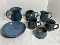 Front view of six assorted pottery pieces including two jugs, two mugs, a plate and small jar all glazed in blue, shown on white background
