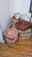 View of stacked natural-tone woven baskets, brown rectangular basket with handle, and tall multicolor basket with lid on wooden floor near wall