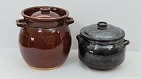 Two ceramic bean pots side by side, taller brown glazed pot and smaller darker glazed pot, both with lids and handles.
