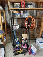 Photo showing the tire rack loaded with various automotive tools including jack stands, trolley jack, tie down straps, jumper cables, and fluid containers on the floor beneath it. Wooden shelves behind with more car tools and supplies.