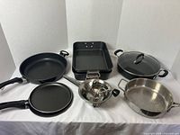 Various cooking implements displayed on a white cloth, including roasting pans, skillets, saucepans, and a food mill, all arranged in front of a white backdrop.