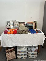Wide view of the crafting lot showing yarn skeins, books, paint containers, blue cases, knitted fabric, and sewing supplies on table with fabric holder stand below