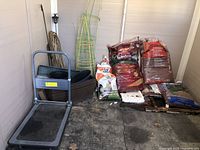 Photo showing push cart replaced with a wheelbarrow, along with tomato cages, planters, and bags of garden materials.