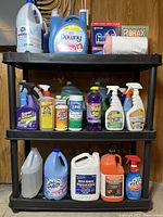 Wide view of household cleaning products organized on black shelving unit. Items include multiple brands and types such as Downy softener, bleach, sprays, and powders.