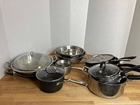 Photo showing assortment of cooking pots and pans with lids, mixing bowls, and colander in varied sizes on a wooden surface.