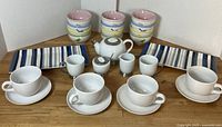 Assorted kitchen dishware laid out on wooden surface including bowls with bird print, striped platters, white tea pot, cream and sugar set, mugs, and saucers