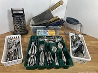 Photo showing three trays with assorted cutlery including forks, spoons, and cutting utensils plus some knives, cleavers, and various kitchen tools behind them on a wooden surface.