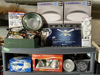 Photo showing entire lot including boxed Homewell flush mount LED lights, Harbor Breeze ceiling fan box, boxed security motion light, flood light fixtures, and assorted light bulbs on shelving.