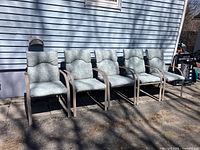 Five metal patio chairs with padded blue floral cushions arranged in a row against an exterior house wall.