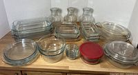 Variety of clear glass bakeware and kitchen storage items including pans, bowls, bottles, and containers arranged on a wooden surface.
