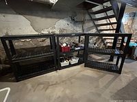 Three black plastic resin shelving units arranged next to each other in basement against wall under stairs.