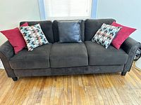 Front view of Lifestyle Solutions dark brown 3-seater sofa with five decorative pillows, placed on hardwood floor in front of window with blinds.