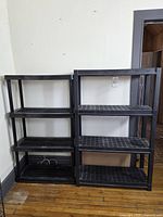 Two black plastic storage shelving units side-by-side on wood floor against white wall.