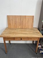 Front view of wooden kitchen island on castors with butcher block placed on top leaning against wall, showing drawer and wheels.