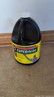 Front-facing photo of black plastic jug with white cap and yellow label showing product name Super Thrive and benefits.