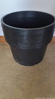 Large 15 gallon black hard plastic plant pot, cylindrical shape with ribbed bands near top edge, standing on floor.
