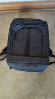 Front view of new black backpack on floor showing front zippered pocket with strap detail and padded shoulder straps.