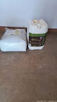 Photo showing the 22.57 kg white bag of Perfect Blend Organics 4-4-4 plant food next to a 5-gallon container of Earthworm Infusion liquid fertilizer on a brown floor against a wall.