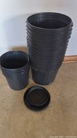 Stack of black plastic soft pots including smaller and larger sizes piled on each other, with some saucers next to the pots on the floor.