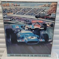 Photo of framed colorful poster showing 1969 Grand Prix race cars on track with crowd stands and shoreline in background.