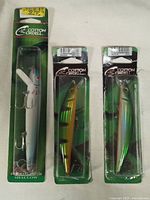 Three Cotton Cordell brand fishing lures in original factory packaging, including one white lure and two green-yellow lures, each with treble hooks.