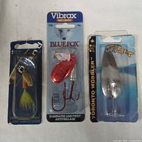 Three factory sealed fishing lures: gold/green spinner with feathered treble hook, red Vibrax Blue Fox spinner, and silver Toronto Wobbler spoon lure.