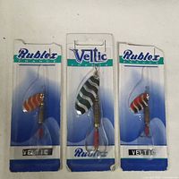 Photo showing three sealed fishing lures in packaging. Two lures have red and gold stripes, labeled Rublex France. Middle lure is larger with black and silver stripes, labeled Veltic France.