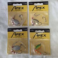 Top view of four sealed Apex fishing accessories packages showing silver spinner and colorful fish design lures