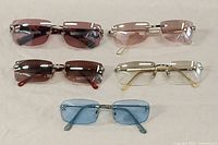Top view of all five colored rimless sunglasses laid flat: two pink, one blue, one red, and one clear light brown lenses.