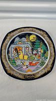 Front of vintage Christmas Carol porcelain plate showing colorful caroling scene with children, animals, house, and gold-trimmed dark blue border.