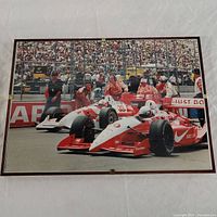 Frontal view of vintage F1 racing photo under glass frame showing red and white race cars with pit crew and audience in background.