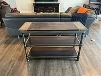 Buffet table with a dark metal frame and three wooden surfaces against a backdrop of a dark leather sectional couch and a TV stand.