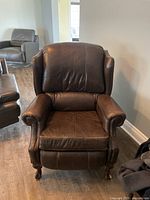 Front-facing image of a brown leather recliner chair with visible creasing and padded armrests. The chair has carved wooden legs and is placed on a wooden floor against a light wall.