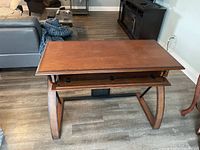 Wooden desk with curved legs and slide-out keyboard tray with two black knobs, medium brown finish, placed on wooden floor in living room setting.