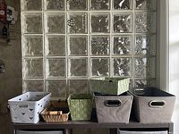Photo showing six storage baskets in different sizes and materials placed on a black surface in front of glass block wall.