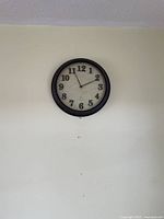 Photo of a round wall clock with a black frame, white face, and black numbers, hanging on a plain wall.