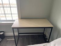 Desk shown with light wood top, black metal legs, placed near window and wall outlet.