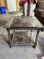 Square end table with wooden top and lower shelf showing dirt and wear, with cylindrical legs and a lower shelf, photographed in a room with various furniture and items.
