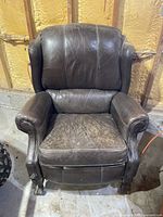 Brown leather recliner chair with heavy signs of wear, some cracking on the leather, cushioned seat, back and armrests, and carved wooden legs.