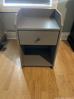 Front view of a modern grey nightstand with one drawer and an open shelf below on hardwood floor under a window.