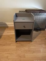 Photo of a gray nightstand with one drawer and a lower open shelf placed on a wooden floor against a light wall.