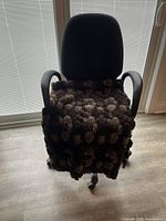 Black office chair with armrests and a brown plush textured cover on the seat, set against a window with blinds.