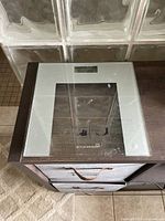 Top-down view of ACCUWEIGH brand glass digital bathroom scale on wooden furniture, showing platform and display area.