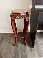 Photo showing a decorative wooden stand with four curved legs, a small lower round shelf, intricate wood carvings under the top, and a pink marble round surface.