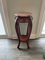 Wooden stand with marble top, visible curved legs and carved floral detail under the marble surface, standing on a wood or laminate floor next to a door.
